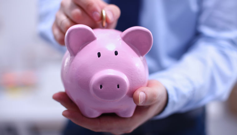 Mann in Business-Dress hält ein pinkes Sparschwein in seiner linken Hand und wirft mit der rechten Hand eine Münze ein.
